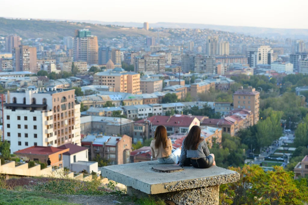 Երկու աղջիկ նստած զրուցում են Երևանի համայնապատկերի դիմաց